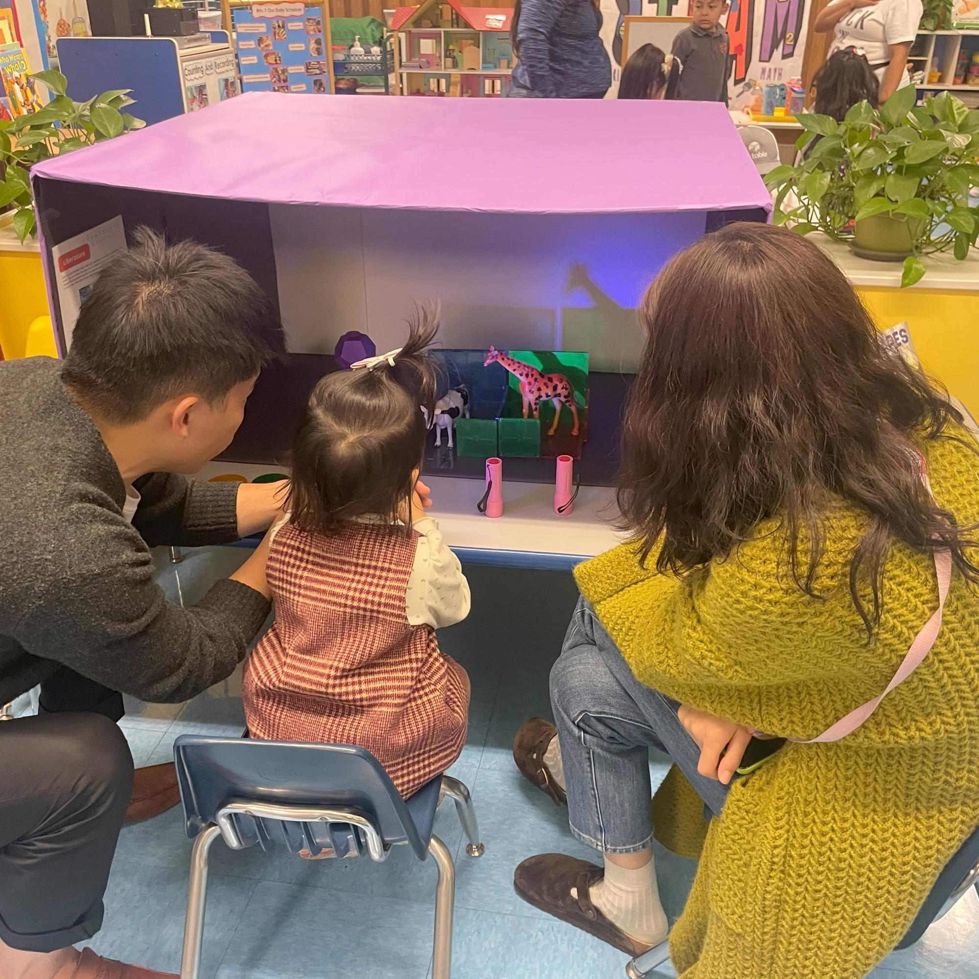 A family observes the shadows made by a light  pointed into a little cardboard theater as part of an interactive STEAM exhibit.