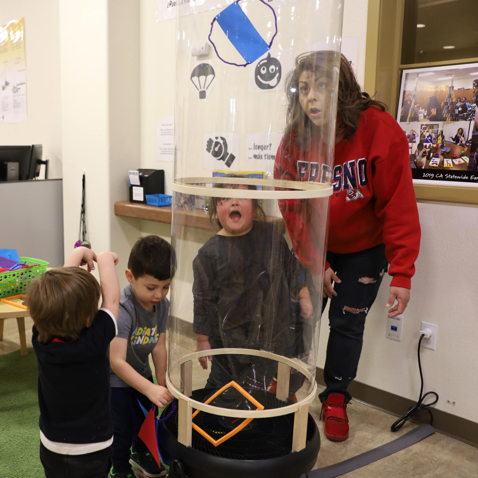 A group of adults and children observe items flying up a wind tube as part of an interactive STEAM exhibit.