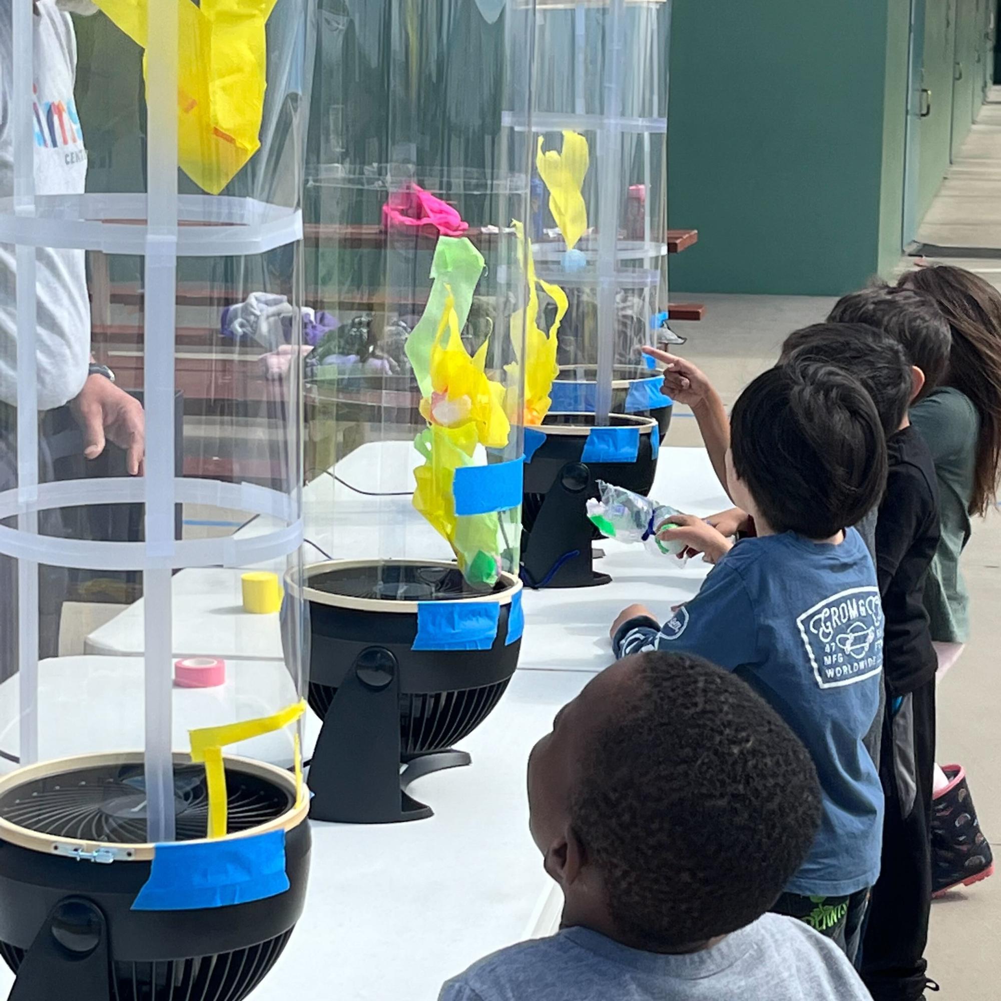 A group of children observe fabric flying up a wind tube as part of an interactive STEAM exhibit.